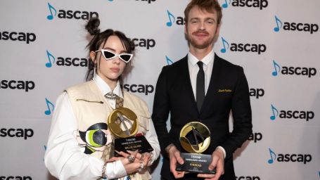 Billie eilish and her brother finneas on the ascap music awards holding 2 different awards, billie wearing a white coat with layered chains and gucci bag while finneas is in a tuxedo 